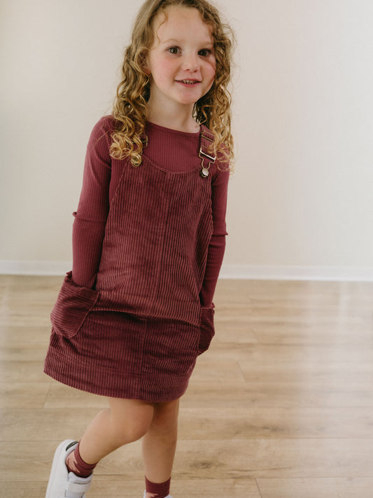 Young girl wearing burgundy corduroy pinafore dress and long-sleeved shirt Corduroy Pinafore Dress - Plum Jam Girls