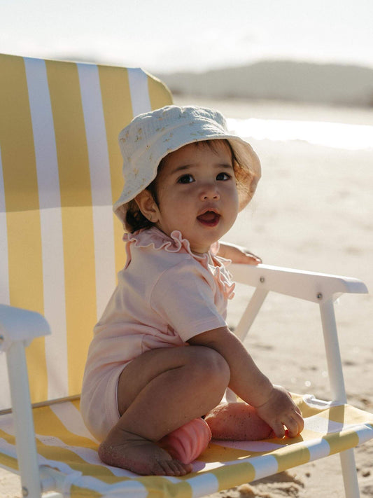 A baby wearing a light pink short-sleeve romper and a white sun hat sits on a yellow and white striped beach chair on a sandy beach.