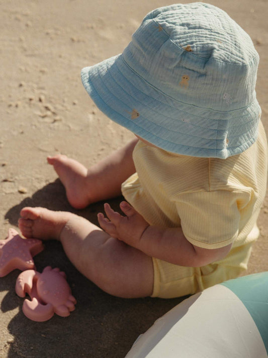 A baby sitting on sand wearing a light blue bucket hat and pale yellow ribbed romper, with pink toy crabs nearby.