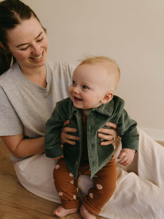Woman holding a baby wearing a green jacket and brown pants in a home setting. Baby Corduroy Jacket - Balsam Baby boy