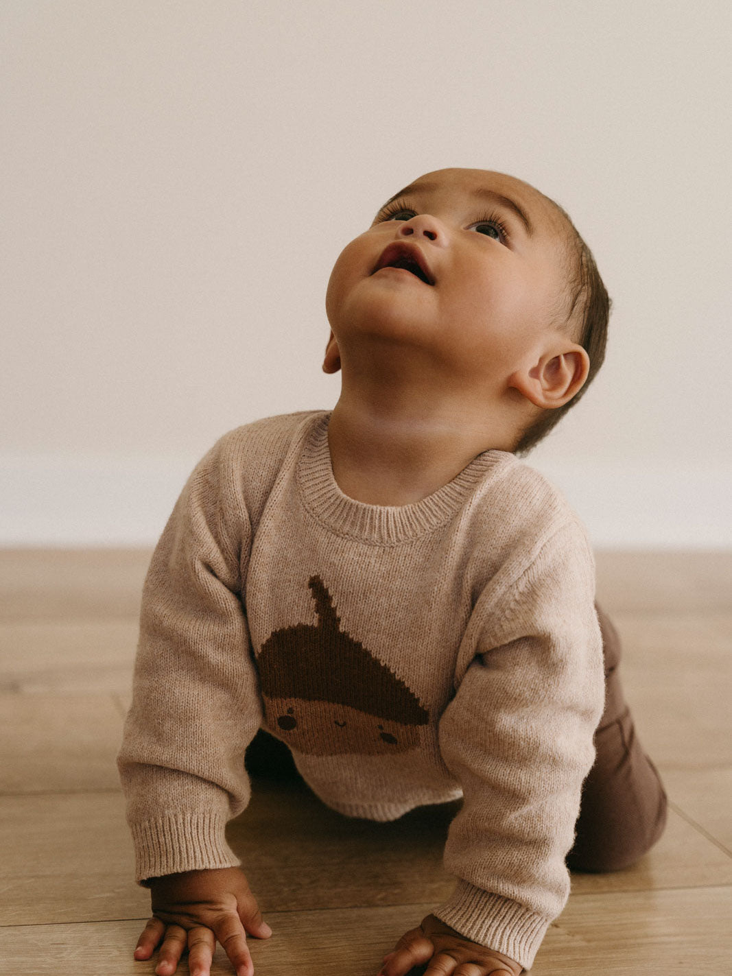 Baby in beige sweater with face design looking upward on wooden surface Baby Feature Print Jumper - Little Acorn Baby boy