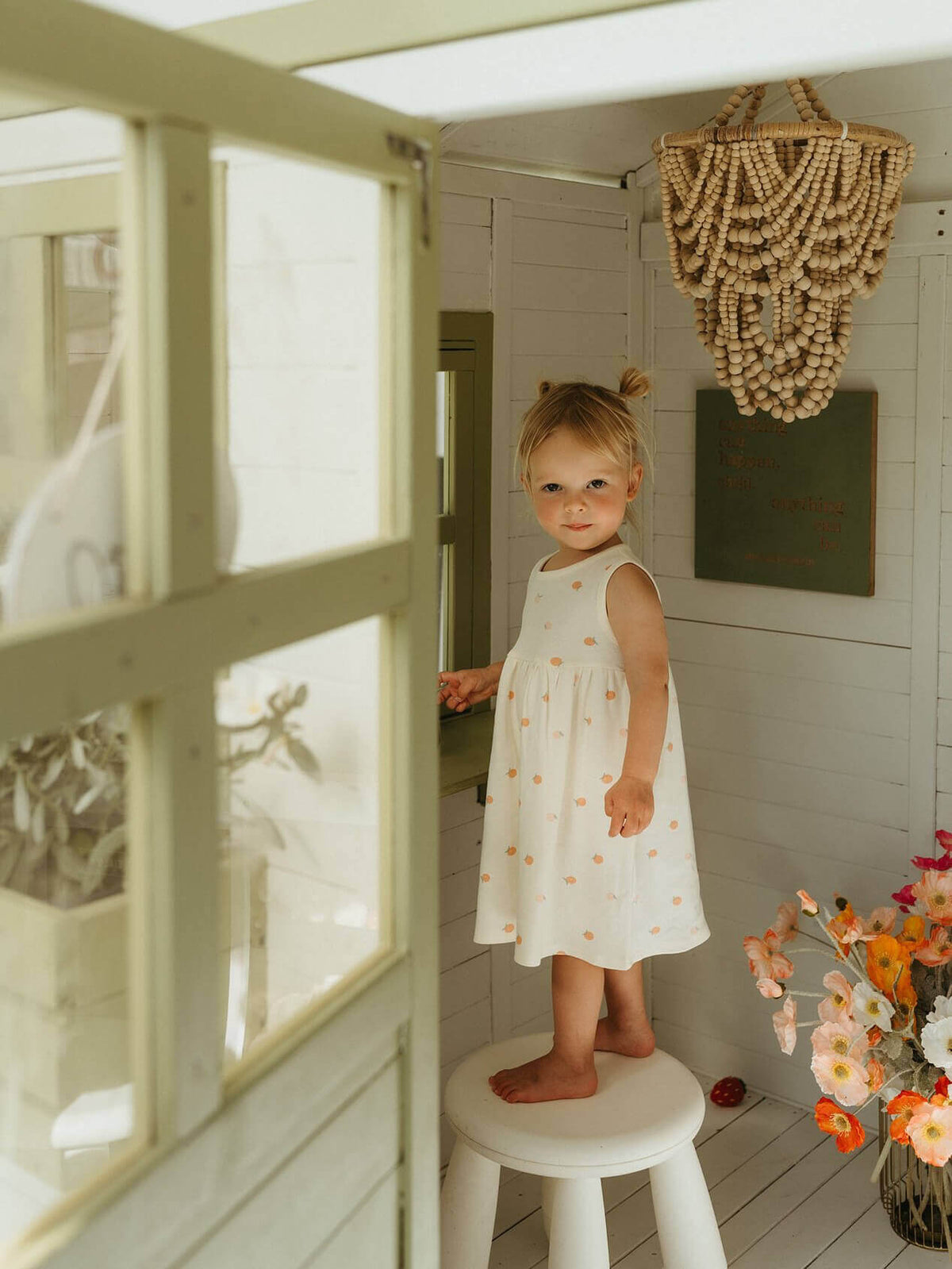 Young girl in a white dress standing on a stool in a bright room with decorative elements. Baby Sleeveless Jersey Dress - Apples Baby girl