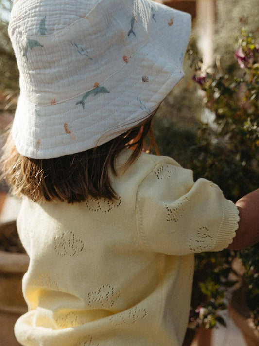 A child seen from behind wearing a cream cotton bucket hat with a small sea creature print and a pale yellow eyelet-knit sweater, standing outdoors near plants.