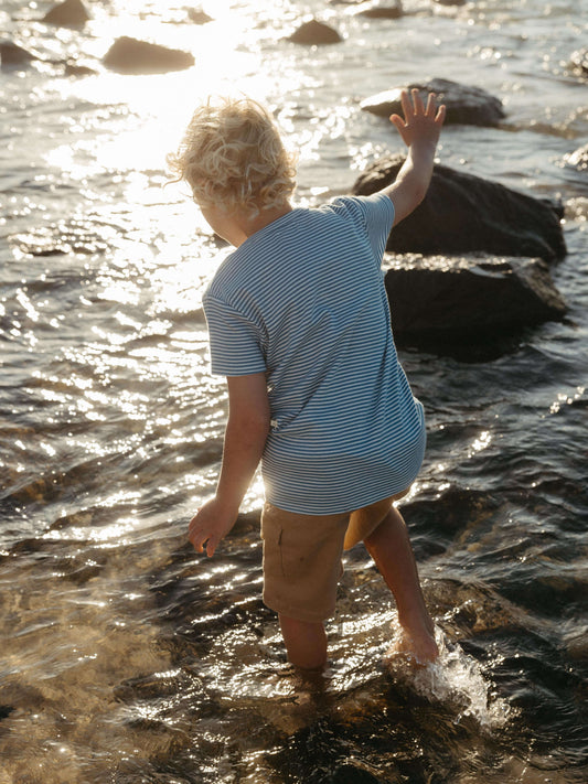 A young child with blonde hair stands in shallow water at a beach or lake, seen from behind, wearing a light blue and white striped t-shirt and tan shorts, with rocks and sparkling sunlight in the water.