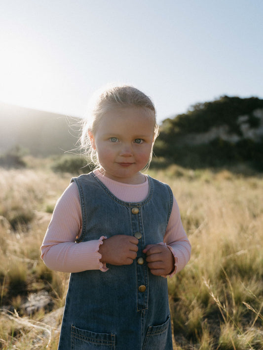 Young girl in denim pinafore dress and pink shirt in grassy field Denim Pinafore Dress - Blue Denim Girls