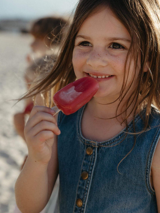 Girl having an ice block at the beach wearing a denim pinafore dress with golden buttons