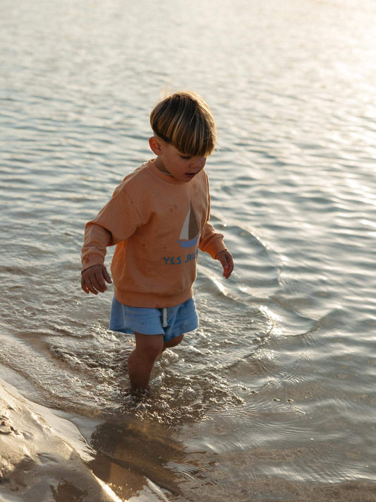 little boy walking on the water at the beach wearing an orange coloured sweatshirt and a blue shorts