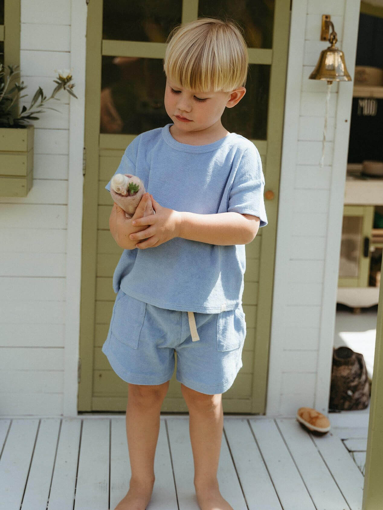 Child in light blue outfit holding a small plant on a wooden deck. Finn Towelling Tee Shirt - Sky Blue Boys