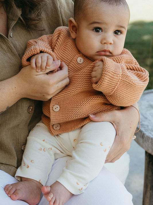 Baby wearing a knitted brown sweater being held by an adult outdoors.