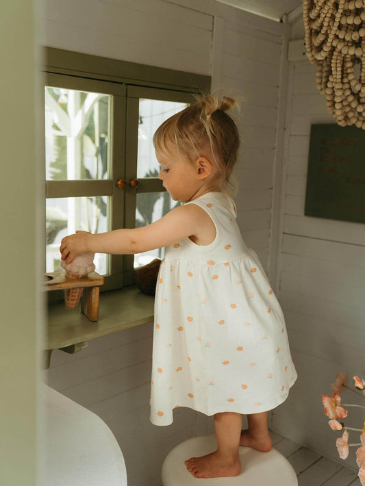 Child in a white dress with orange apples standing on a stool inside a playhouse.