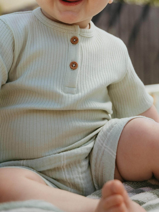 Child wearing a light-colored ribbed shirt and shorts sitting outdoors.