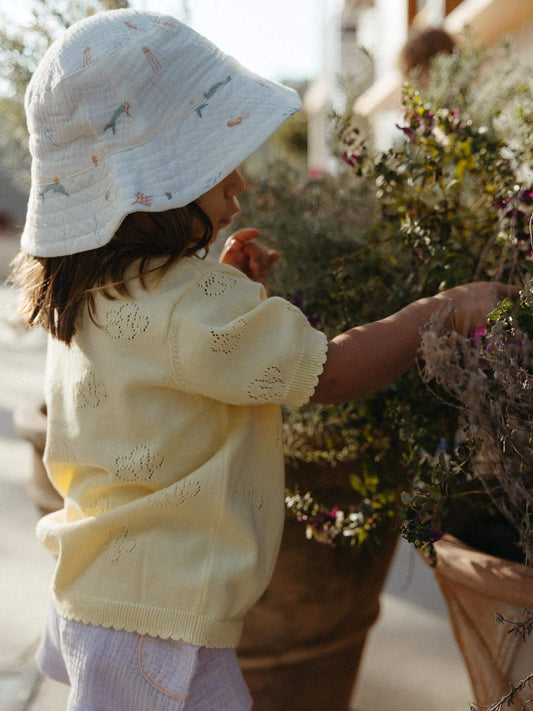 A toddler wearing a pale yellow knit cardigan and a white bucket hat with small colorful bird prints, standing outdoors next to potted plants.