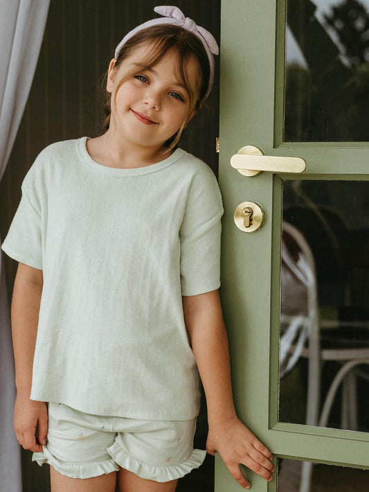 Young girl in a light green outfit standing next to a green door. Lara Towelling Tee Shirt - Mint Girls