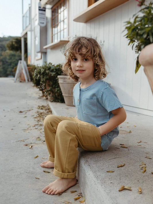A young child with curly light brown hair sits barefoot on a concrete ledge outdoors, wearing a blue and white striped short-sleeve henley shirt and mustard yellow pants.