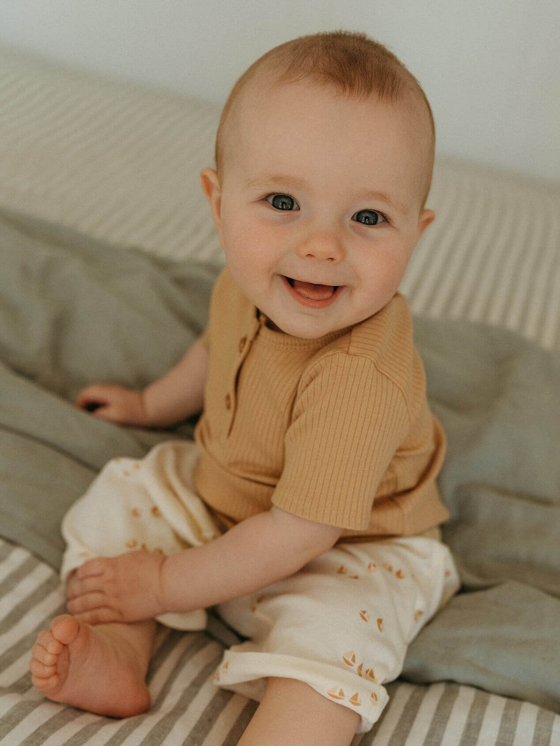Smiling baby sitting on a bed in a tan tee and Sailing print shorts.