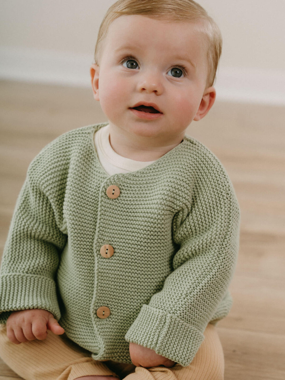 Baby wearing a sage knitted cardigan with wooden buttons and cream trousers.