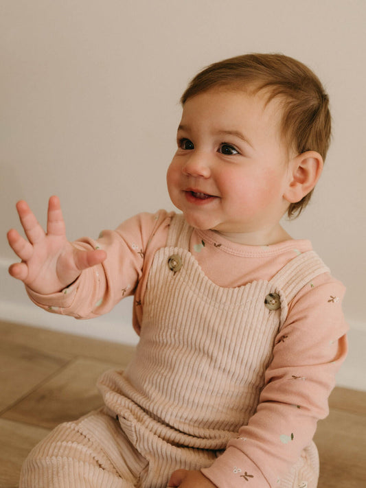 Baby sitting in Yes Mila corduroy overalls in Clay layered over a printed long sleeve tee