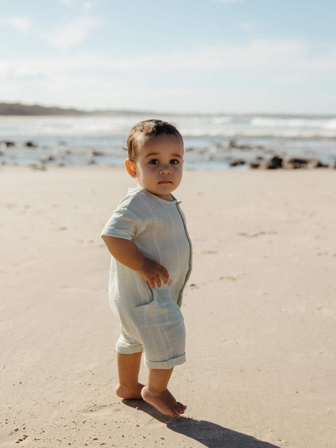 Baby on a New Zealand beach in Yes Mila organic muslin baby clothes, showing the open muslin weave