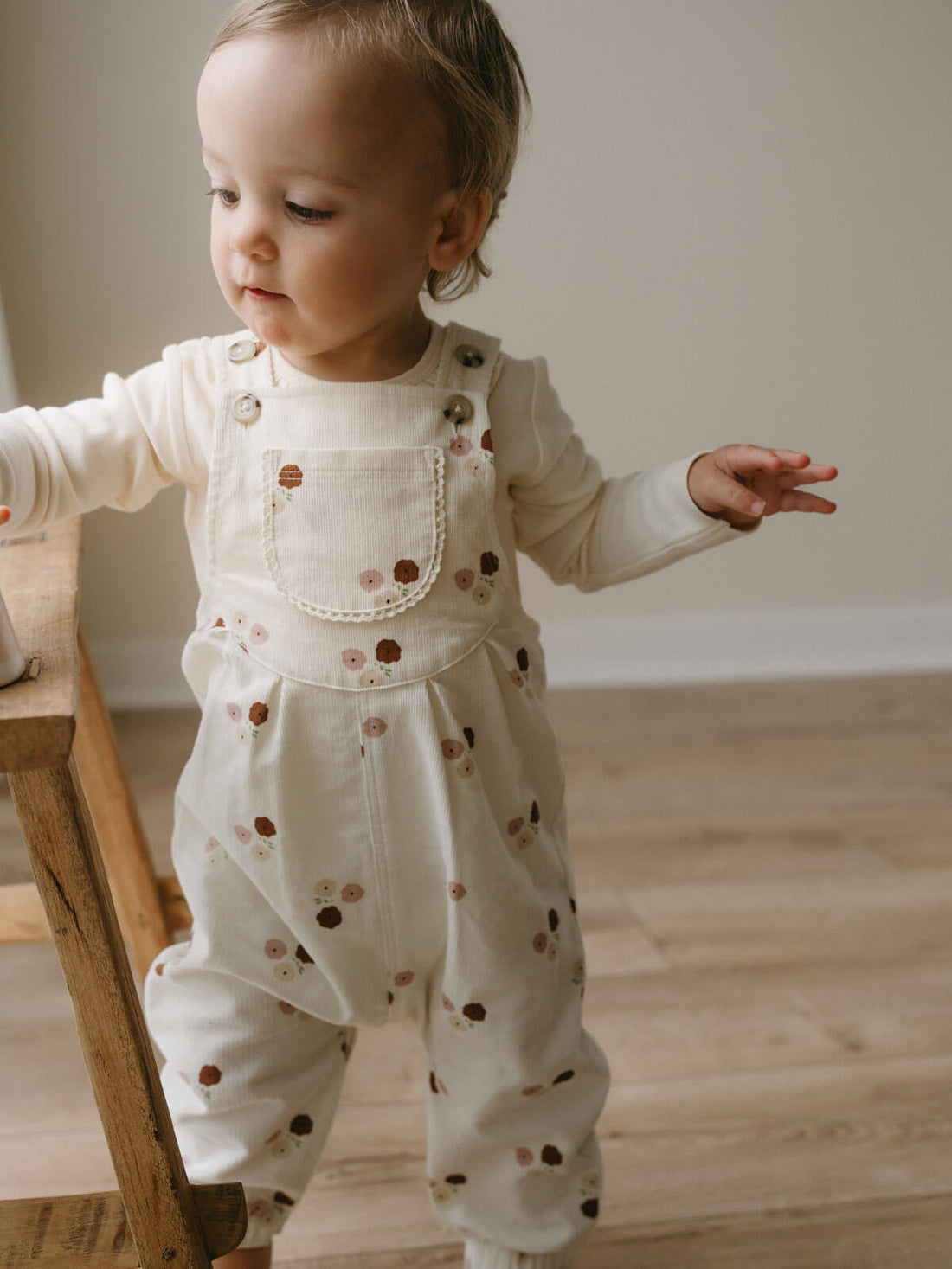 Baby standing beside a chair wearing floral Yes Mila overalls over a long-sleeved top.