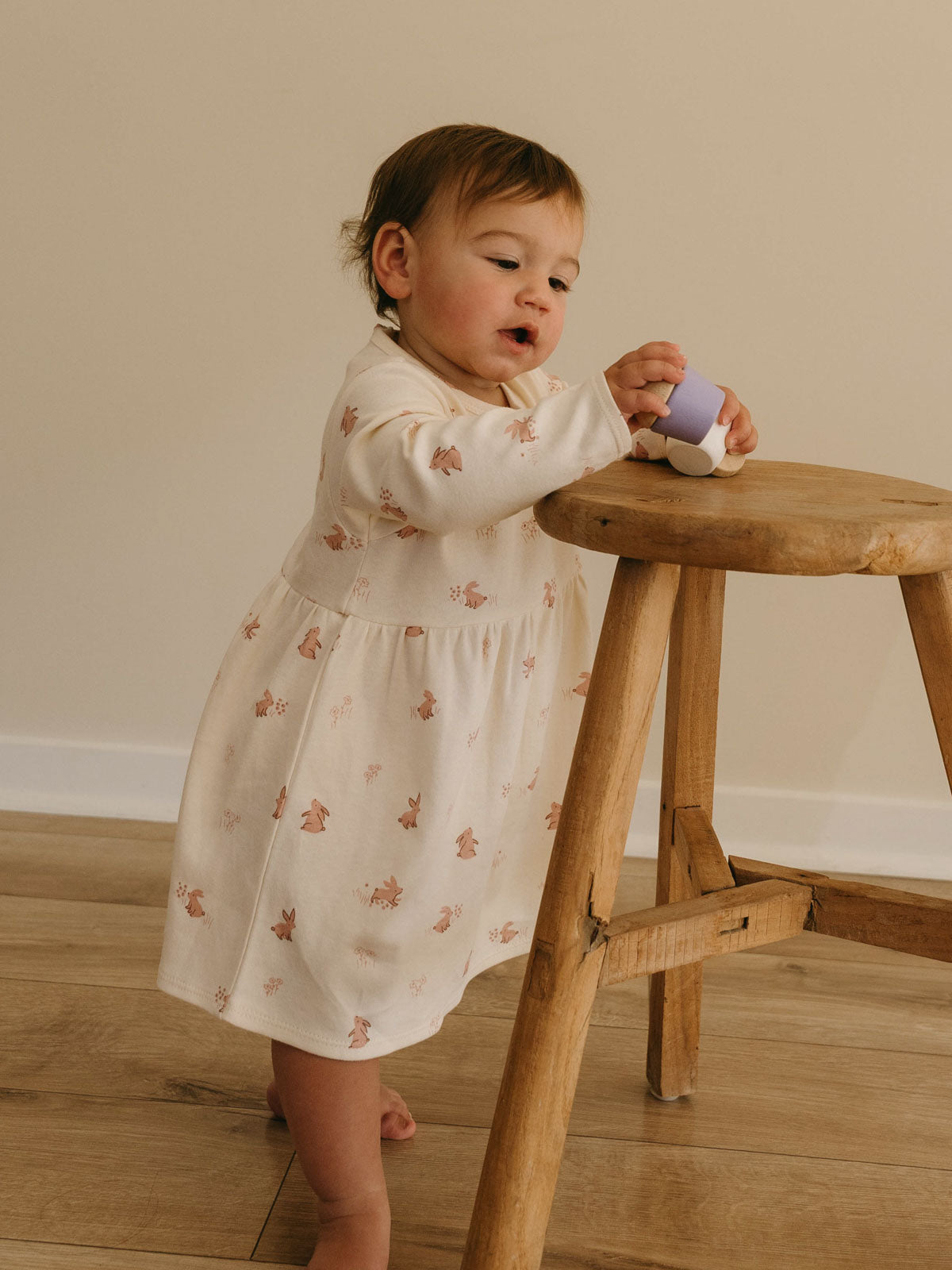 Toddler in a white bunny-print dress standing beside a wooden stool holding a toy Baby Long Sleeve Jersey Dress - Meadow Friends Ecru Baby girl