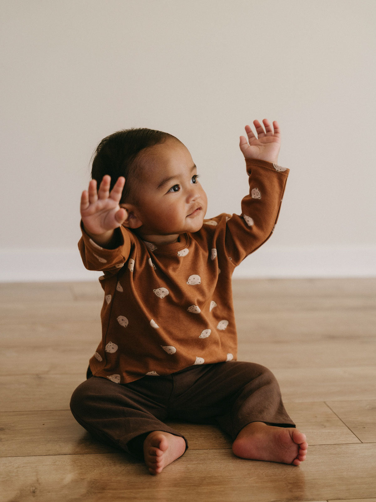 Young child sitting on wooden floor wearing rust-colored printed long sleeve top Baby Printed Long Sleeve Tee - Cosy Bear Russet Baby boy