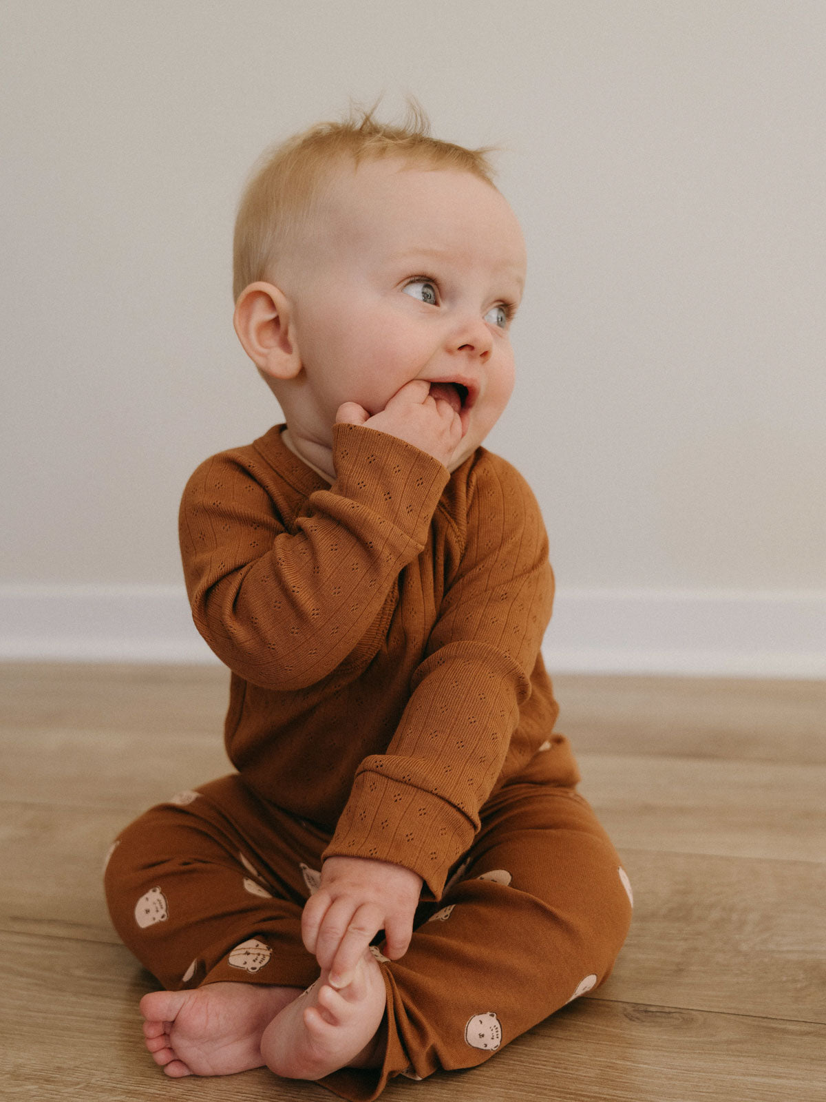 Baby in rust-colored outfit sitting on wooden floor with hand near mouth Pointelle Crossover Bodysuit - Russet Baby boy