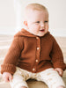 Baby wearing a rust-colored ribbed knit cardigan with buttons, sitting on wooden surface Ribbed Hooded Cardigan - Russet Baby boy