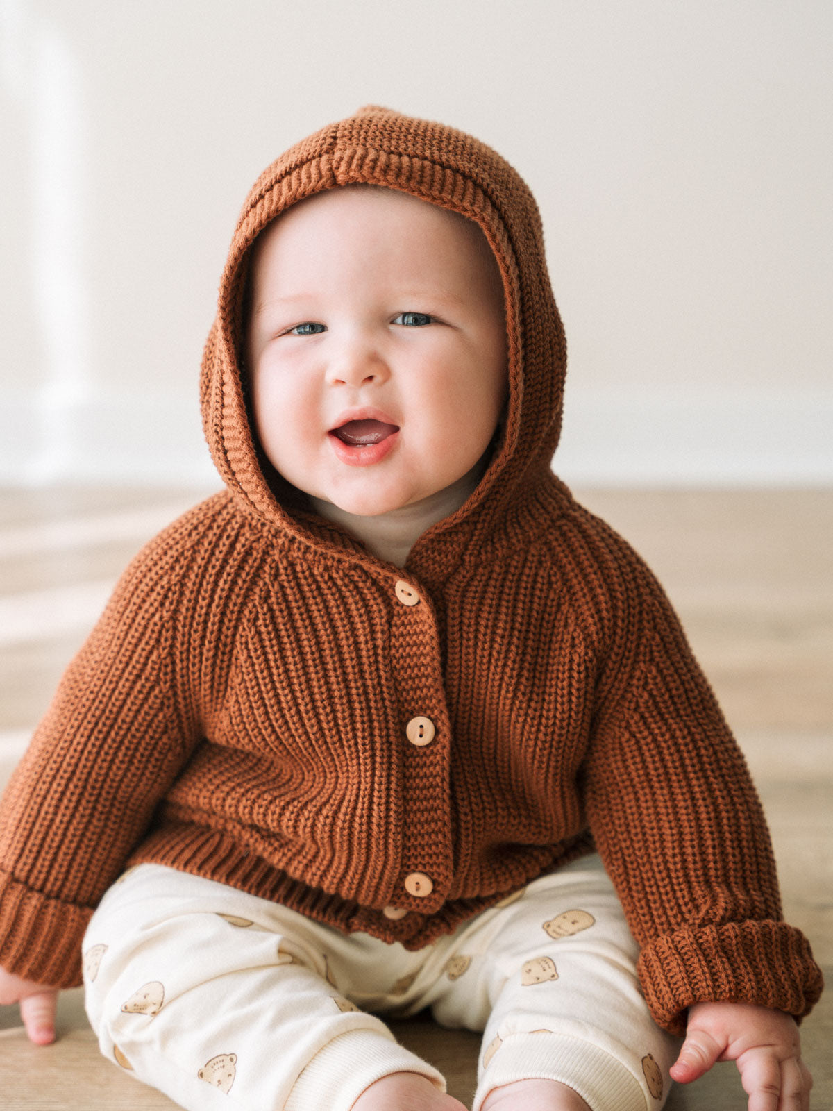 Baby wearing a rust-colored knitted hooded cardigan with wooden buttons sitting down Ribbed Hooded Cardigan - Russet Baby boy