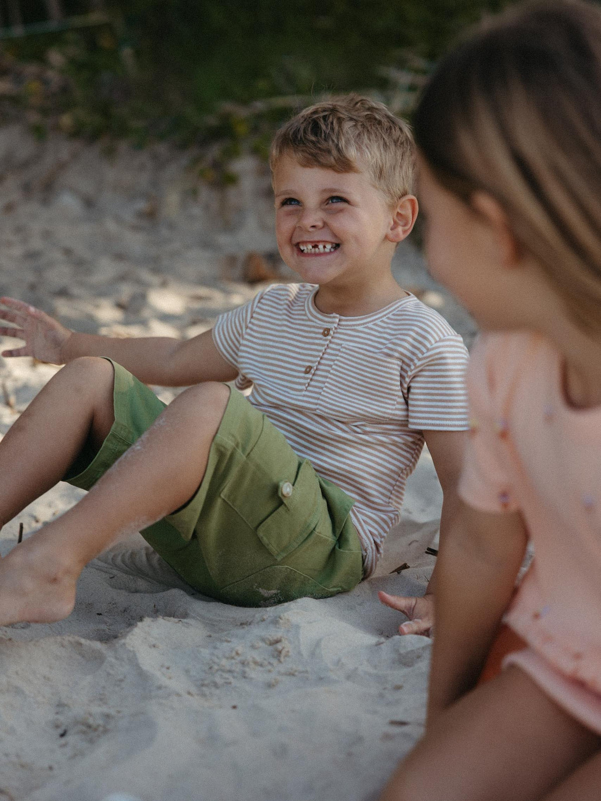 A young boy sitting on sand at the beach, smiling and looking to the side. He is wearing a white t-shirt with thin beige horizontal stripes and olive green shorts.
