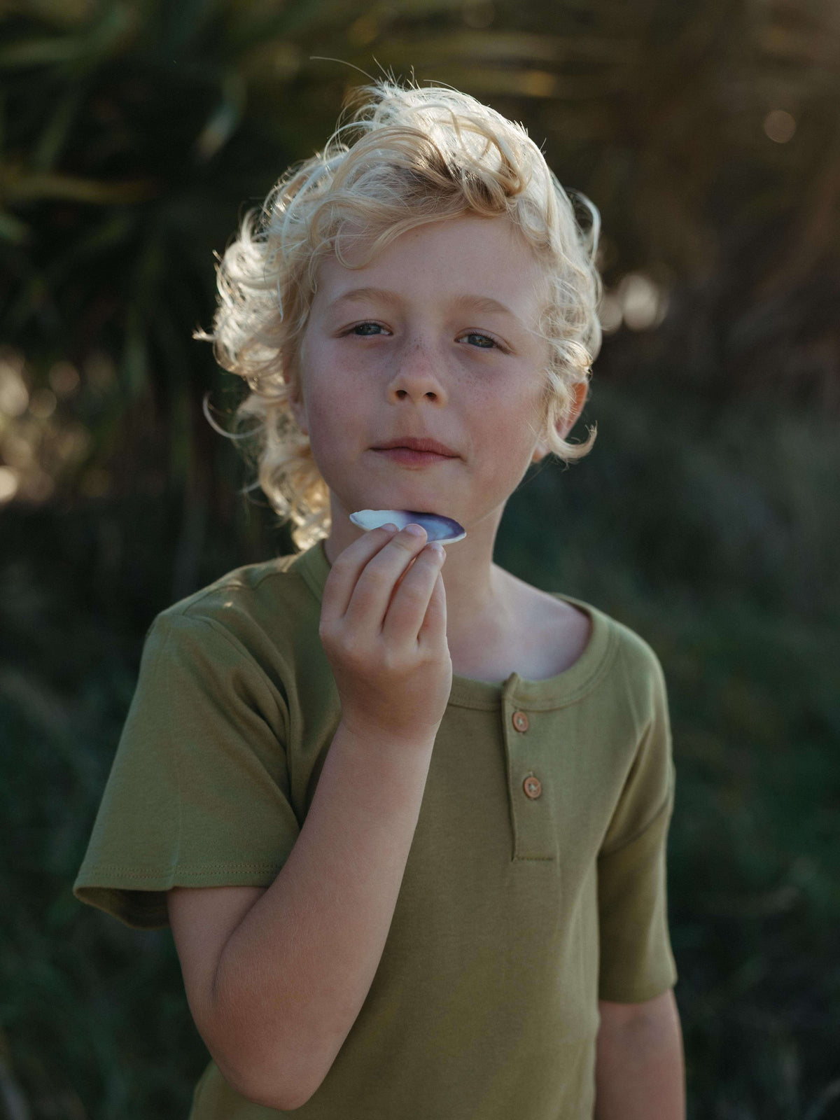 Boy holding a shell at the beach wearing a green tee