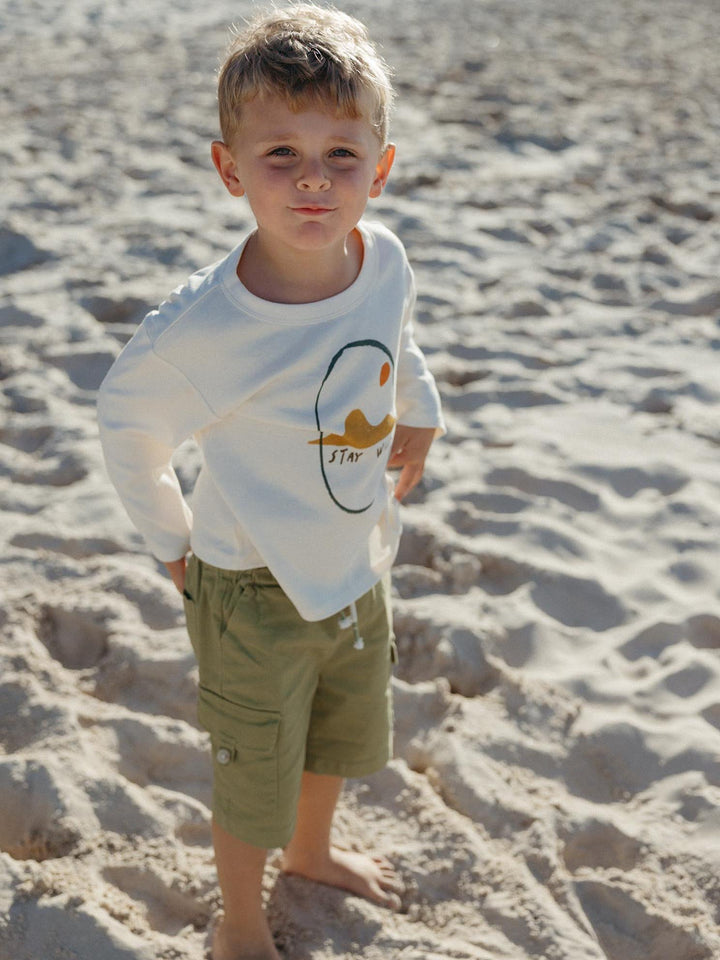 A young boy standing on a sandy beach wearing a white long-sleeve t-shirt with a graphic print and olive green cargo shorts.