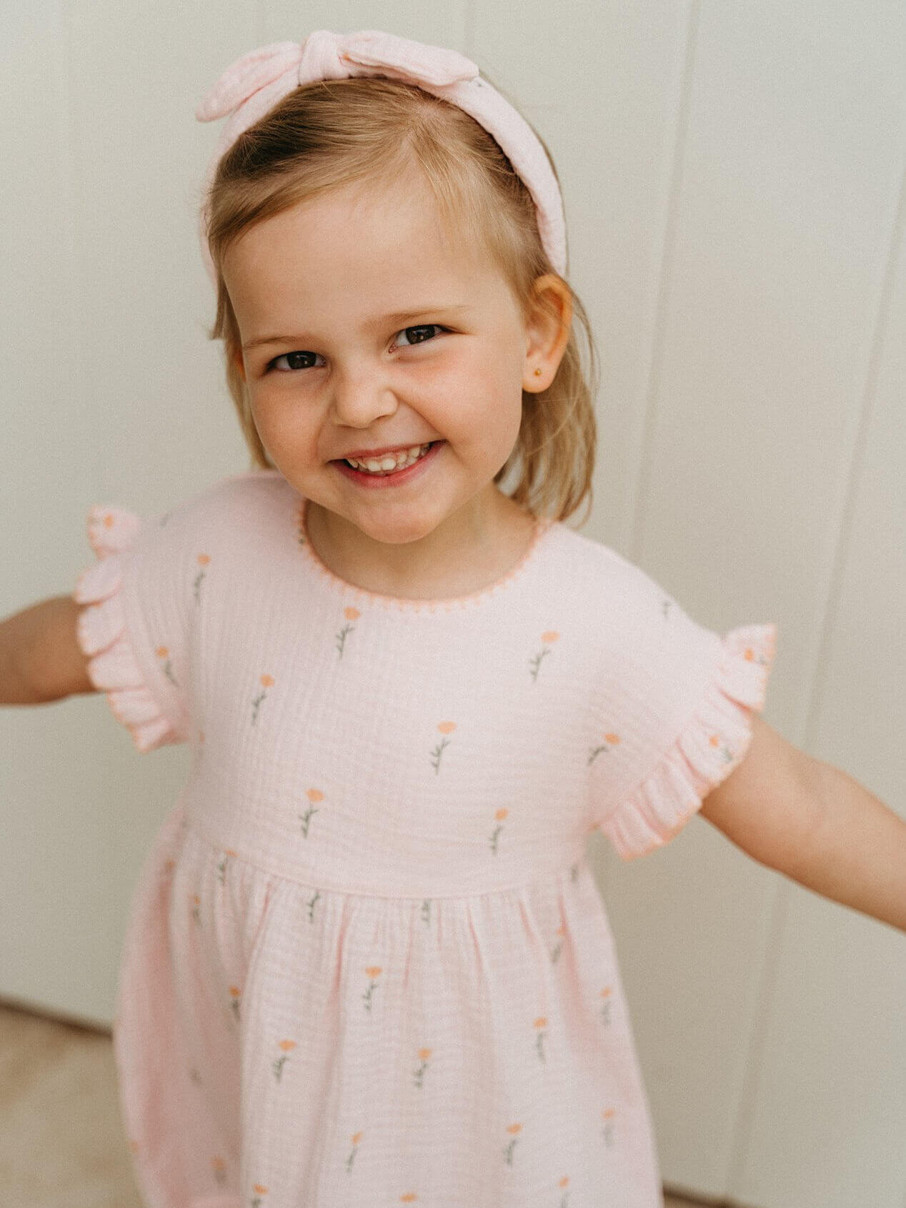 Young girl wearing a pink dress with ruffled sleeves and a matching headband, standing against a light background.