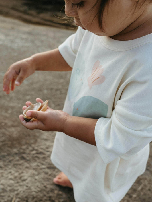 Child holding a shell on a sandy beach wearing a white shirt with nature-themed design.