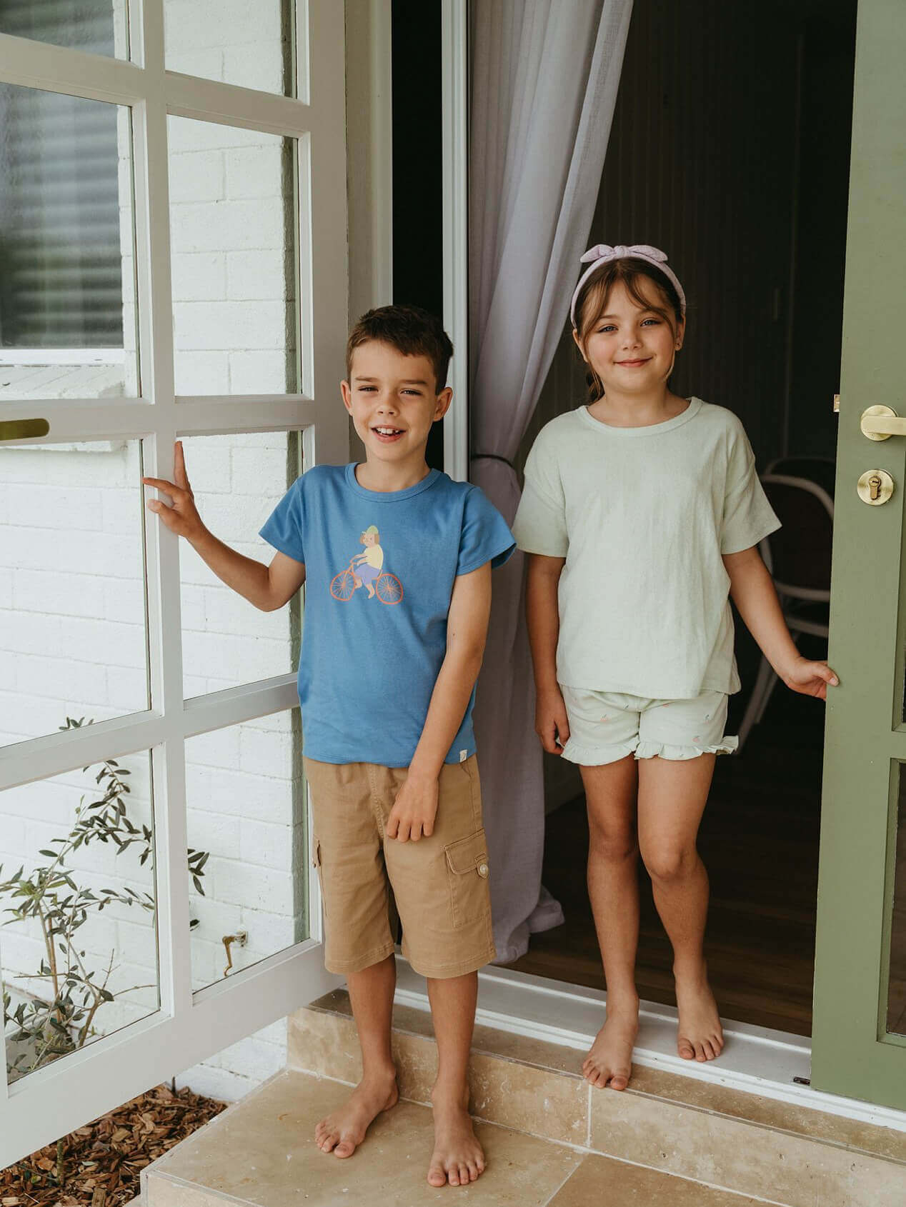 Two children standing on a porch with a white door and window in the background. Axel Short Sleeve Tee - Milo Biking Boys