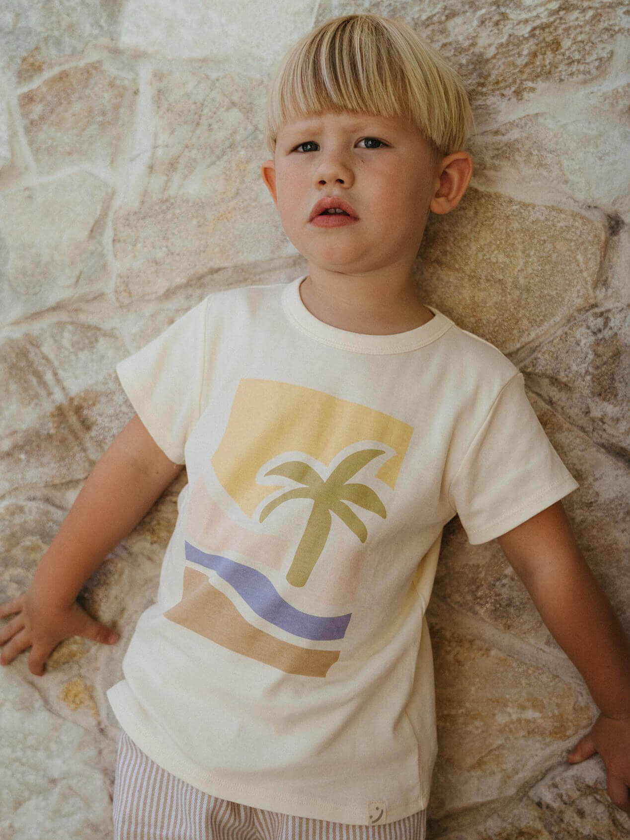 Child wearing a t-shirt with a palm tree design against a stone wall. Axel Short Sleeve Tee - Palm Beach Boys