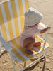 A baby wearing a light pink outfit and a white sun hat sits on a yellow and white striped beach chair on sandy ground.