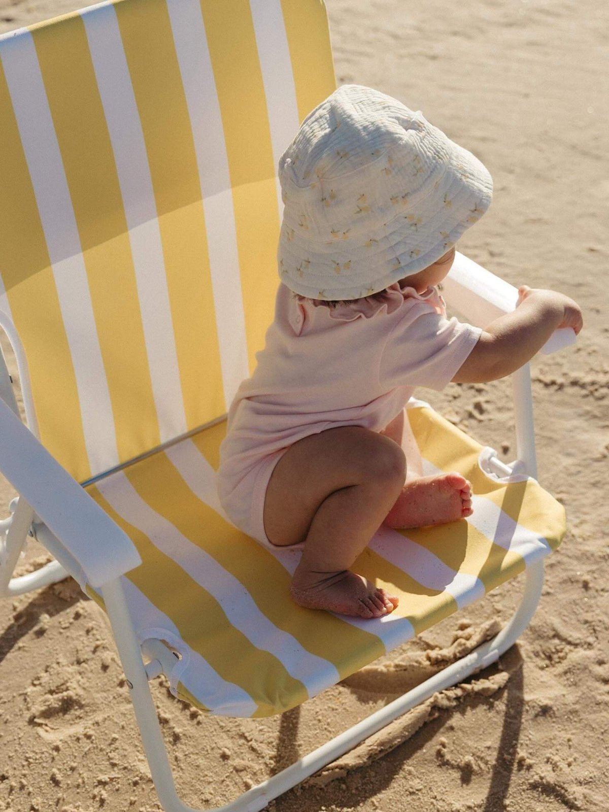A baby wearing a light pink outfit and a white sun hat sits on a yellow and white striped beach chair on sandy ground.