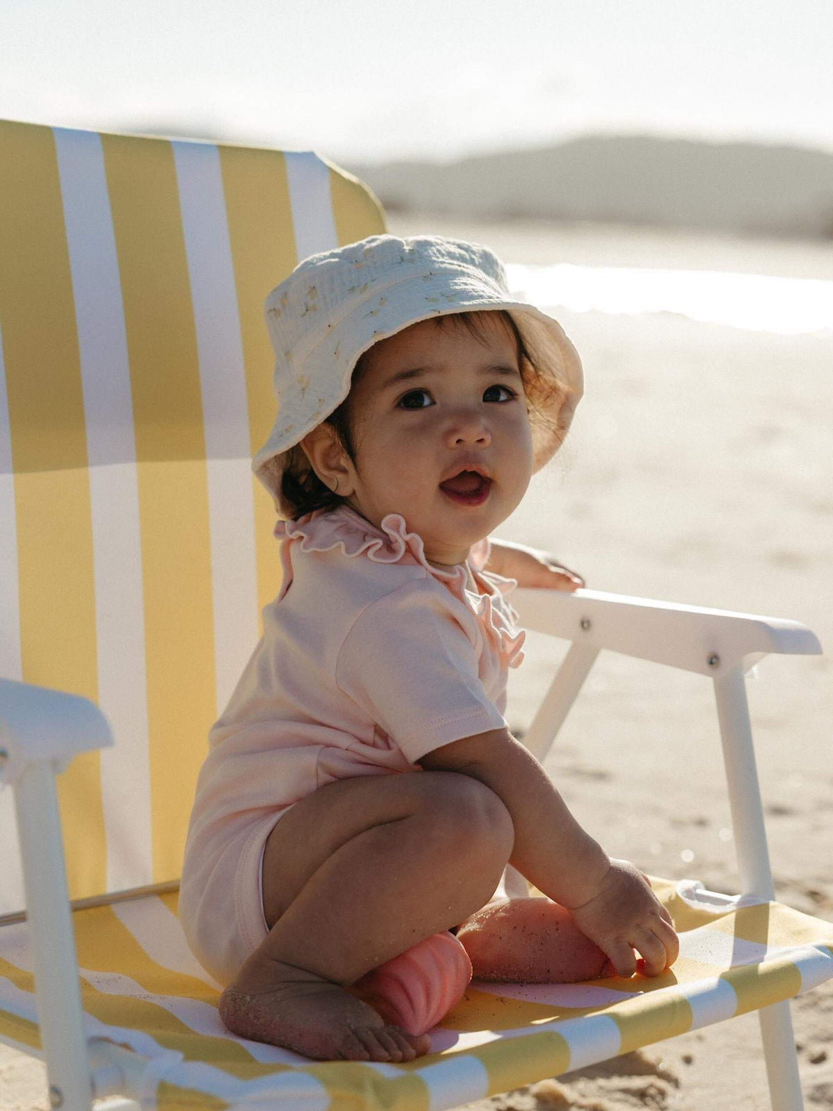 A baby wearing a light pink short-sleeve romper and a white sun hat sits on a yellow and white striped beach chair on a sandy beach.