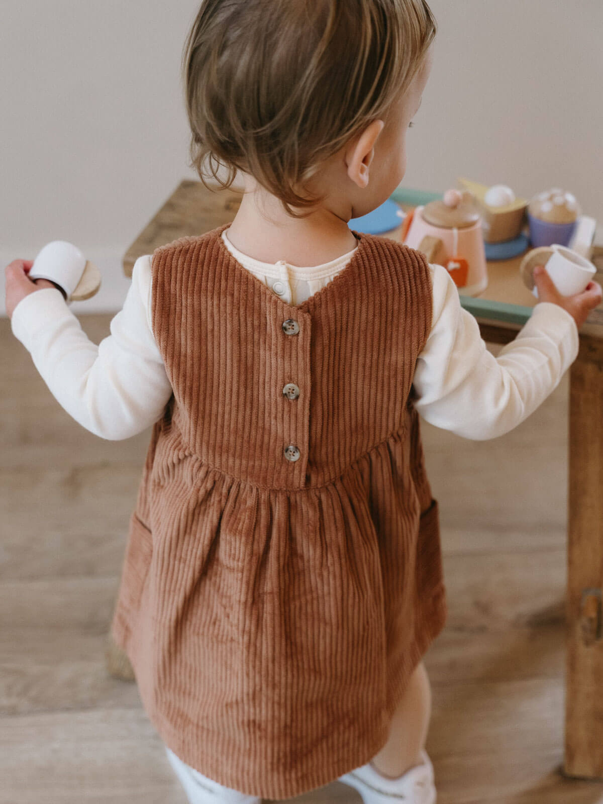 Child wearing a brown corduroy dress with a white shirt underneath, standing in front of a wooden table. Baby Corduroy Dress - Russet Baby girl