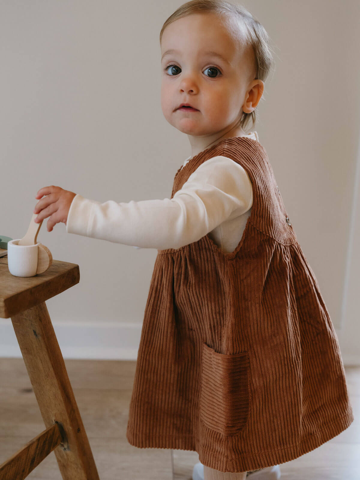 Child wearing a brown corduroy dress with a white shirt underneath, standing next to a wooden stool. Baby Corduroy Dress - Russet Baby girl