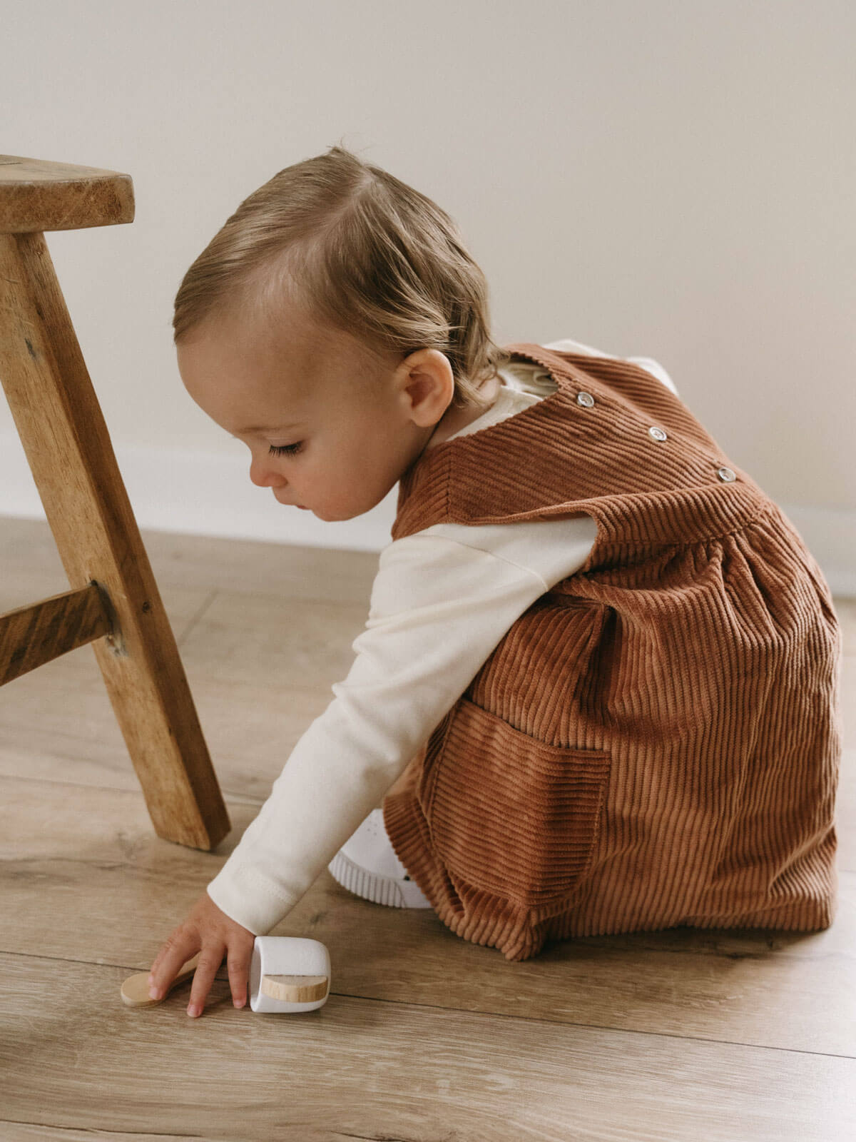 Child in a brown corduroy dress sitting on a wooden floor. Baby Corduroy Dress - Russet Baby girl