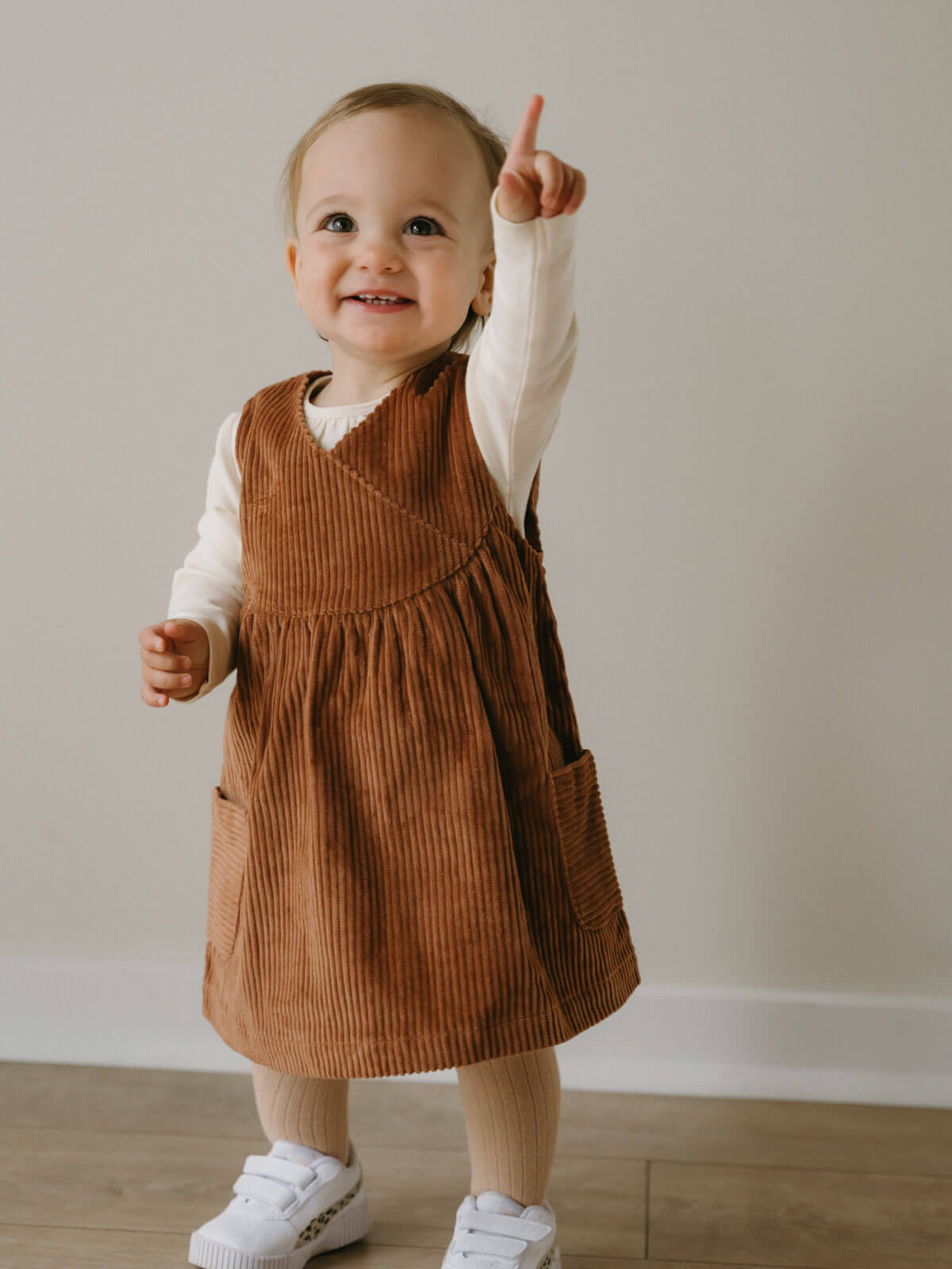 Child wearing a brown corduroy dress with a white shirt underneath, standing on a wooden floor. Baby Corduroy Dress - Russet Baby girl