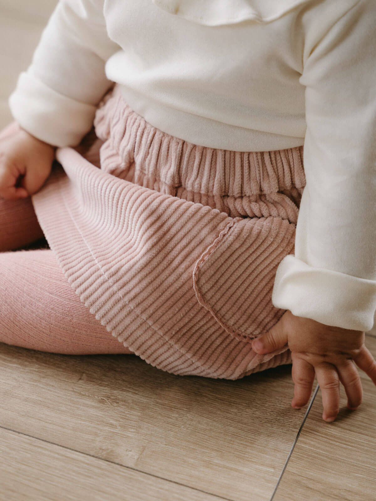 Person wearing a white top and pink ribbed pants sitting on a wooden floor. Baby Corduroy Skirt - Blush Baby girl