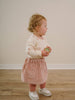 Child wearing a pink skirt and white sneakers holding a green object indoors. Baby Corduroy Skirt - Blush Baby girl