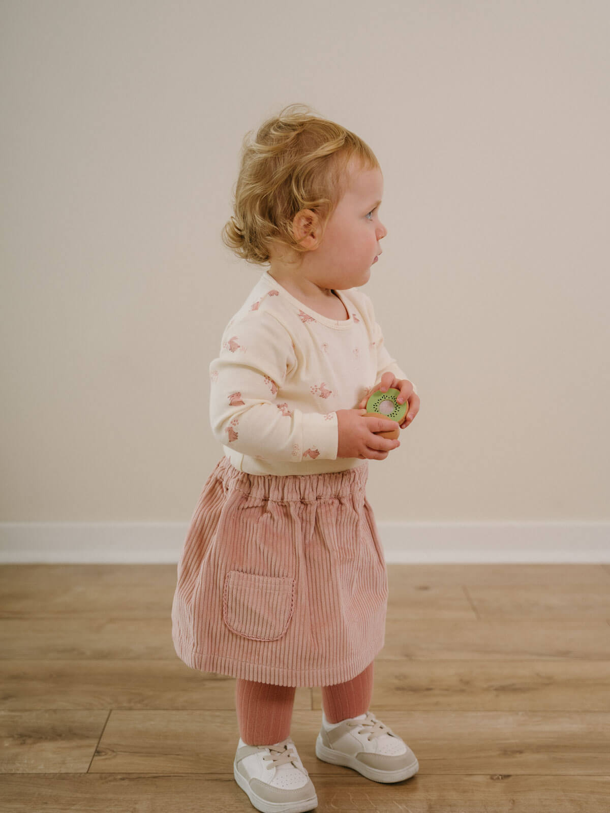 Child wearing a pink skirt and white sneakers holding a green object indoors. Baby Corduroy Skirt - Blush Baby girl