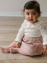 Child wearing a white top with ruffled collar and pink skirt sitting on a wooden floor. Baby Corduroy Skirt - Blush Baby girl