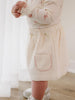 Girl wearing a cream-colored corduroy apron with a pocket, standing indoors. Baby Corduroy Skirt - Ecru Baby girl
