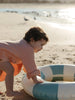 A toddler in a pink knit top and orange skirt crawls on a sandy beach next to a blue and white striped inflatable pool float.