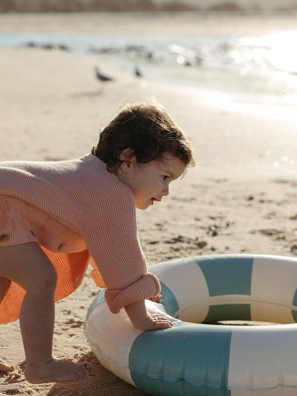 A toddler in a pink knit top and orange skirt crawls on a sandy beach next to a blue and white striped inflatable pool float.