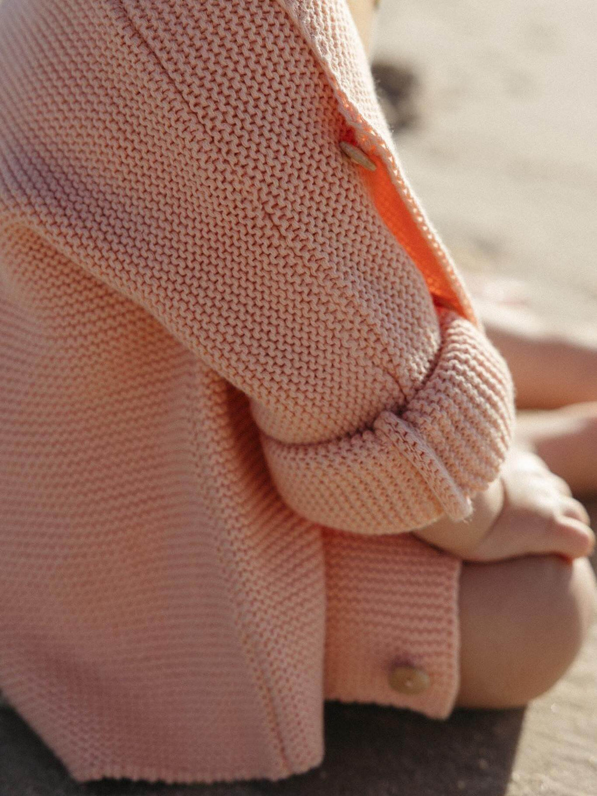 A close-up of a child wearing a soft peach knit cardigan, with the sleeve rolled up and a small wooden button visible, in an outdoor setting.